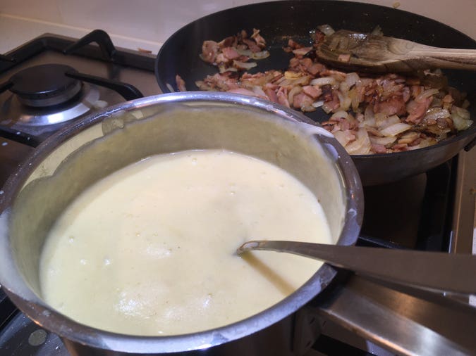 adding cheddar cheese and cayenne pepper to béchamel, plus onion and bacon frying in the background