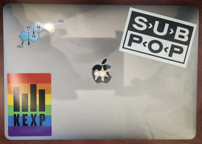 a Macbook Air with four stickers. In a corner each is a "sub pop" record label sticker and a pride "kexp" (seattle radio station) in the other. Almost in a corner is a foil estrogen molecule and directly covering the apple logo is a foil caffeine molecule.