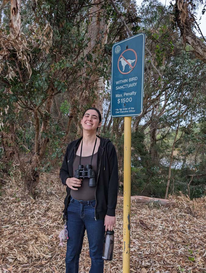 doggirl standing in front of a "[no dogs symbol] in bird sanctuary" sign with a shit-eating grin