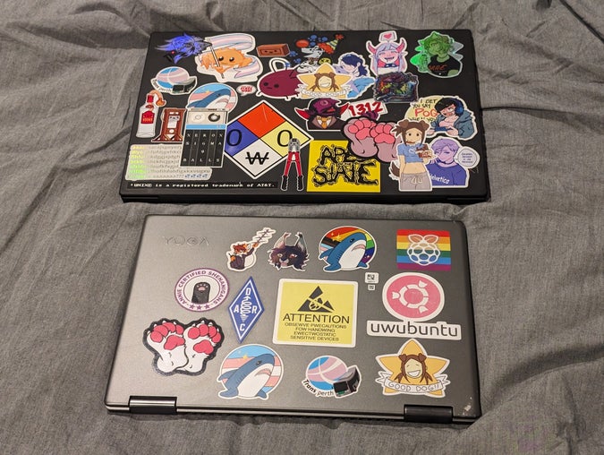 two laptops on a bed, with a variety of pretty gay stickers. one has stickers like a doggirl eating chocolate, an nfpa hazard diamond that reads “0W0”, and a keysmash that reads “aaaarsarajsgoyery[…]fziyxouv aaaaaaa??? 💦💦💦💦”, while the other has a trans flag blåhaj sticker, some ham radio stickers, and a sticker with a crossed out paw that reads “ATTENTION: OBSEWVE PWECAUTIONS FOW HANDWING EWECTWOSTATIC SENSITIVE DEVICES”.